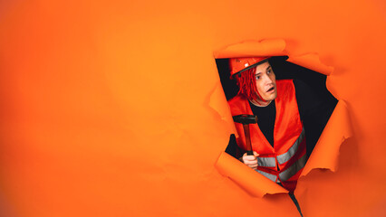 Shocked male construction worker in overalls with hammer looking out of hole of orange background. Young man looking around in state of shock about what he did.