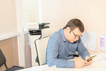 Obraz premium Man employee in blue shirt and glasses sitting at the desk in office with smartphone using digital technology applications for business development. Business, busy working day concept
