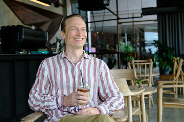 A man sits in a cafe and drinks a drink. The man is holding a drink. The man smiles.