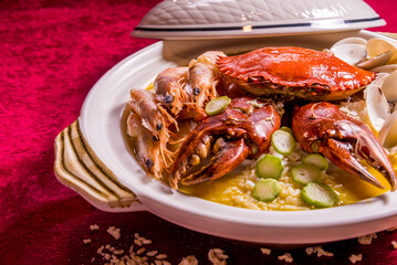 stewed mixed seafood platter crab, shell clam, tiger prawn in hot clay pot with rice and yellow chicken stock congee on red background chinese banquet menu