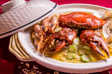 stewed mixed seafood platter crab, shell clam, tiger prawn in hot clay pot with rice and yellow chicken stock congee on red background chinese banquet menu