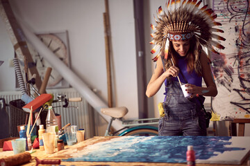 A young female artist with a war bonnet in the studio is mixing and preparing the paint for a new painting. Art, painting, studio - obrazy, fototapety, plakaty