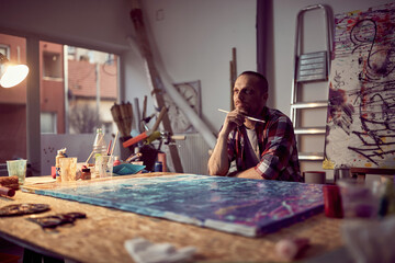 A young male artist is brainstorming while working on a new painting in the studio. Art, painting, studio - obrazy, fototapety, plakaty