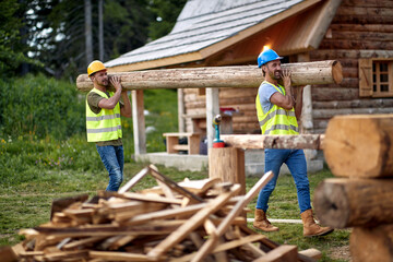Two young male builders carry timber at cottage construction site in the forest. Construction,...