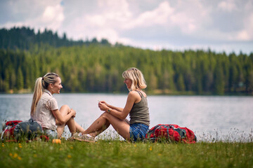 Two woman sitting in grass in front of lake and talking with backpacks by their side. Leirsure time. Trip, weekend, fun, lifestyle, nature concept