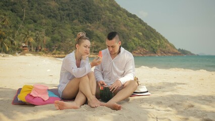 Fototapeta premium The cheerful love couple holding and eating slices of watermelon on tropical sand beach sea. Romantic lovers two people caucasian spend summer weekend in outdoor. Hat, backpack white shirt beachwear.
