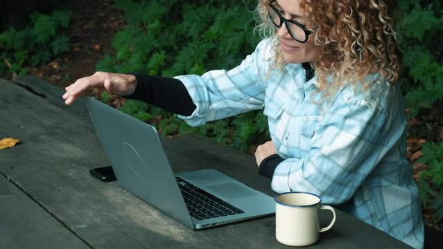 Happy modern adult woman working on laptop computer outdoor sitting on a wooden bench with forest nature around. Concept of remote worker and digital nomad freedom lifestyle