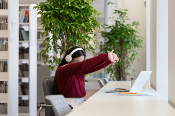 Young student girl stretching arm while working on laptop in university library. Asian woman tired from exam preparation wear headphones watching online webinars and lectures at distant education