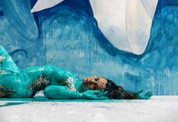 Side view of a turquoise green and white abstract painted sexy nude woman, lying on the floor in the blue studio, abstract body art painting