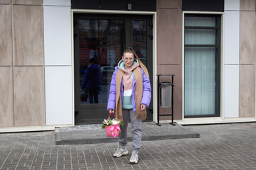 A young attractive girl in a stylish opposing suit and a bouquet of flowers walks through the streets of the city.