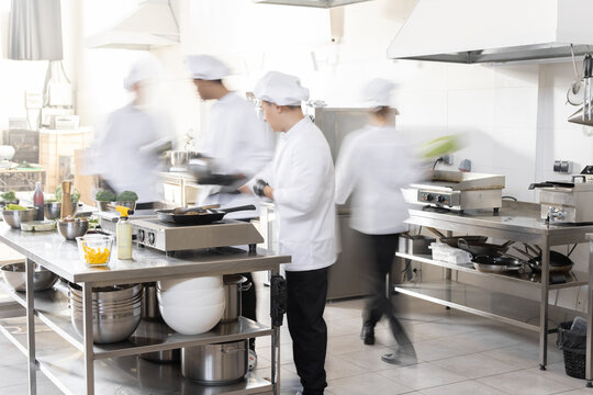 Chef Cooks Working In Professional Kitchen. Chefs Hurry Up, Actively Cooking Meals For Restaurant. Long Exposure With Motion Blurred Figures