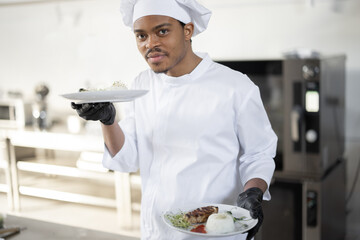 Portrait of young handsome smiling chef in uniform standing with ready meal in the kitchen. Happy...