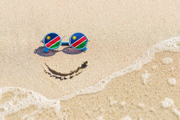 Fototapeta na wymiar A painted smile on the sand and sunglasses with the flag of the Namibia. The concept of a positive and successful holiday in the resort of the Namibia.