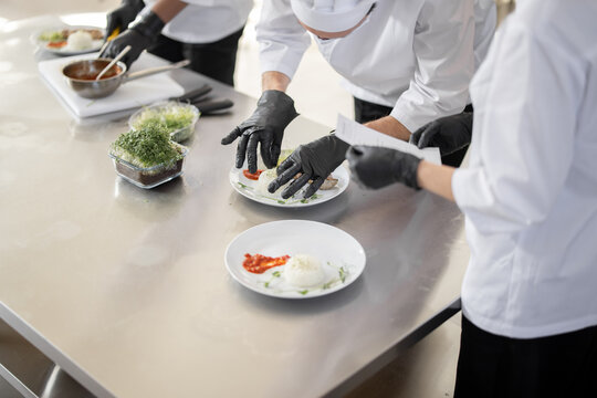 Cooks finishing main courses, putting greens on plates with rise and meat, close-up on table with meals in the kitchen. Chefs wearing gloves and uniform. Concept of haute cuisine and professional
