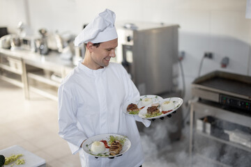 Young handsome smiling chef in uniform standing with ready meals in the kitchen. Happy caucasian cook working at restaurant. Concept of professional occupation and haute cuisine