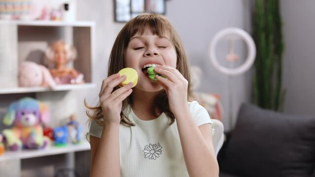 Attractive smiling cute preschool kid girl holds two tasty cookies in her hands and makes her choice, then bite macaron in green color. Traditional French multicolored macaroon. Food concept.
