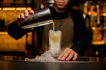 Fototapeta na wymiar front view of glass full of ice pieces in which the hand of bartender pours a cocktail from shaker