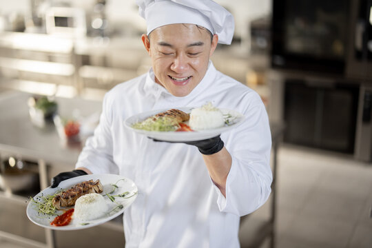 Portrait of young happy handsome smiling chef in uniform standing with ready meal in the kitchen. Happy asian cook working at restaurant. Concept of professional occupation and haute cuisine