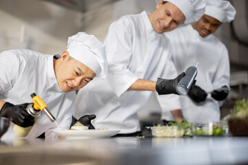 Multiracial group of cooks finishing main courses while working together in the kitchen. Asian chef bakes a dish with a burner, other guys serving dish on background. Team prepares meals for the