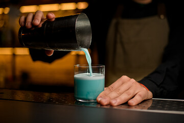 Fototapeta na wymiar front view of glass in which the male hand of bartender pours a blue cocktail from a shaker