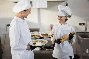 Chef standing with ready meals, talking with female cook in the kitchen. Happy caucasian cook working at restaurant. Concept of professional occupation and teamwork