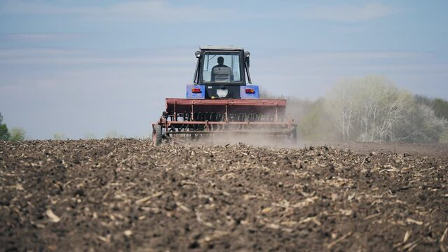 Rear view of a farmer on a tractor sowing grains of corn in a dry agricultural field. Dust follows the agricultural machine. Agribusiness in the spring