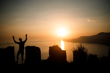 Fototapeta premium A woman or a man standing on a rock and looking directly at the sunset by the sea. The concept of nature and beauty. Orange sunset. Silhouette at sunset