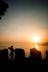 A woman or a man standing on a rock and looking directly at the sunset by the sea. The concept of nature and beauty. Orange sunset. Silhouette at sunset