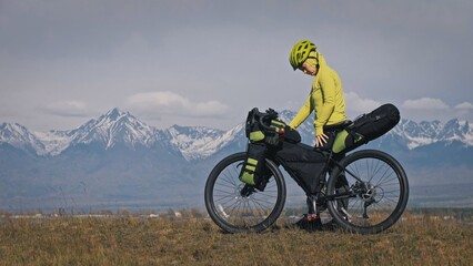 The woman travel on mixed terrain cycle touring with bikepacking. The traveler journey with bicycle bags. Sport tourism bikepacking, bike, sportswear in green black colors. Mountain snow capped.