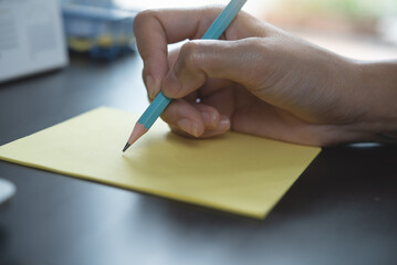 Close up of business woman hand with pencil writing on an adhesive note on table in office, focus on hand and pencil, work planning, to do list, ideas notice reminder