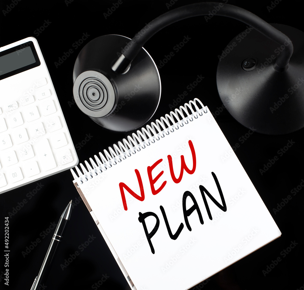 Poster opened paper notebook with text new plan with a pen, calculator and table lamp on black background.