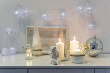 The concept of decorating the house for Christmas. Lighted candles, a garland, a disco ball, an angel figurine on a white chest of drawers. Place for text