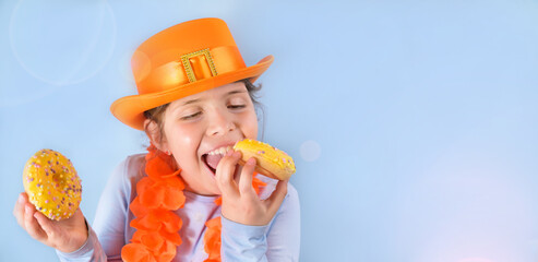 King's Day in Holland, traditional festival on April 27 in the Netherlands. Little girl in a festive orange hat on a blue background eats colored donuts. Copy space. Banner