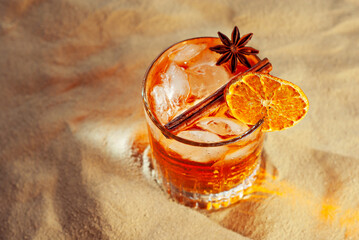 Orange cocktail on yellow sand. Alcoholic drink with ice, orange and cinnamon close up.