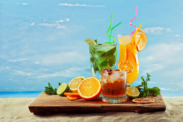 Orange and green cocktails on a wooden board. Alcoholic drink with ice, orange, mint and lime on the sand. Sunset, sea and sky in the background. A mixture of different drinks with fruit.