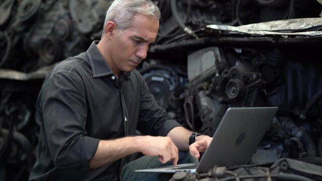 senior engineer using laptop computer checking list old used car engine in machinery parts shop warehouse. old technician man Repair vehicle automobile machine in Recycle motor factory