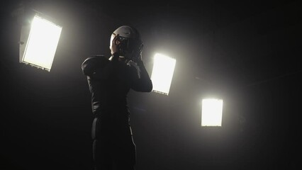 Portrait of a bearded American football player putting on helmet and preparing for winning game. Confident man in uniform into backlit dark stadium. Masculine footballer. Close up. Slow motion.
