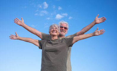 Happy senior couple standing with open arms with blue sky background. Beautiful smiling white-haired seniors enjoying retirement and freedom on an outdoor excursion