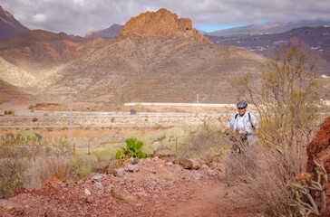 Cheerful senior woman riding in a country road with bike, cyclist wearing sport helmet. Mountains and cloudy sky in background