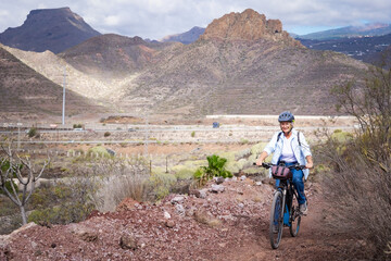 Obraz premium Cheerful senior woman riding in a country road with bike, cyclist wearing sport helmet. Mountains and cloudy sky in background