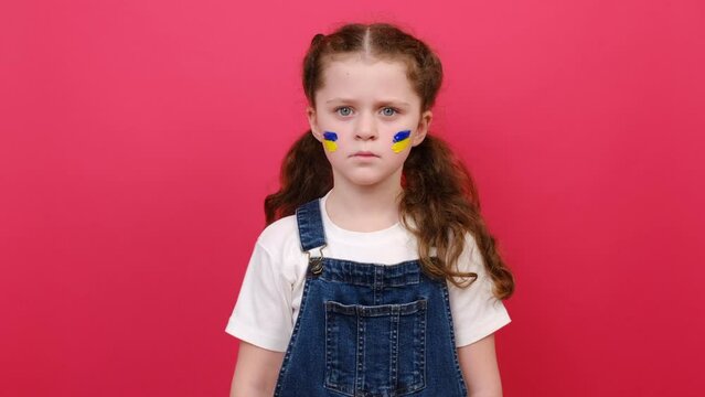 Portrait of upset little child girl with flag of ukraine on face, serious looking at camera, isolated on red background. Stop russian aggression. Evacuation of civilians. Freedom to Ukraine, Crisis