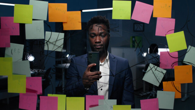 African-American businessman use smartphone working on glass board late in office