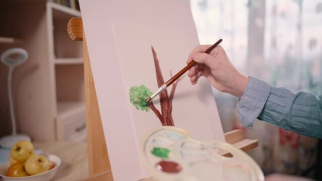 Girl paints picture of tree with paints on paper with brush at home. Close-up of picture. Slider shot.