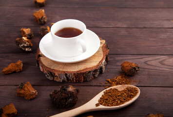 Chaga mushroom coffee drink in a white cup on a wooden stand, scattered pieces of chaga and a spoon with chaga powder on a wooden background. Copy spaes.