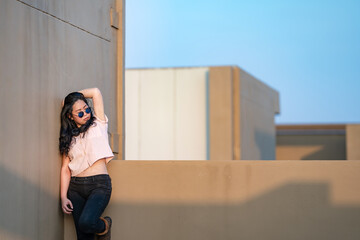 A cute Asian woman stands up and raise elbow up against the wall and wears blue sunglasses at the rooftop of the building at sunset time.