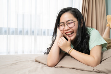 Beautiful glasses Asian woman is laughing while lying on her stomach on the bed in bedroom with a pastel green - brown color theme.