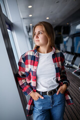 A beautiful attractive ukrainian woman with blond hair is standing by a large window, wearing momom jeans and a t-shirt with a plaid shirt. Girl in casual comfortable clothes