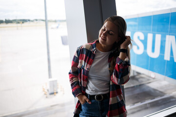 Obraz premium A beautiful attractive ukrainian woman with blond hair is standing by a large window, wearing momom jeans and a t-shirt with a plaid shirt. Girl in casual comfortable clothes