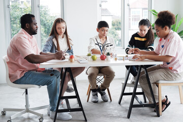 Young African teacher or tutor is sitting on chair and teaching drawing and painting with watercolor of art for Caucasian students or pupils in classroom at school. Education for artist Concept.