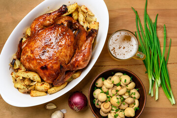fried chicken meat and roasted potatoes on a white platter, a glass of beer, champignon mushrooms and green onions, delicious food on a wooden background